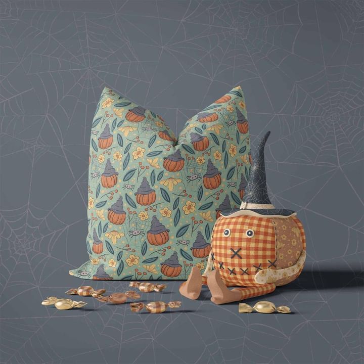 Halloween pillow mockup with a pumpkin pattern next to a plush toy pumpkin wearing a witch hat on a gray surface.