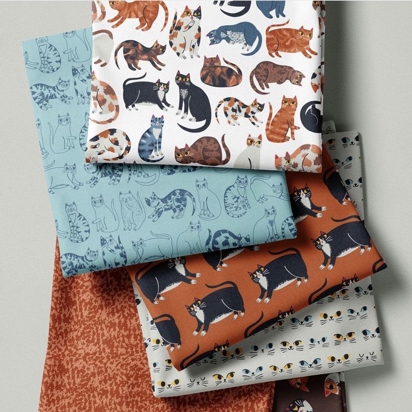 Fabric stack mockup showing six folded fabrics with various cat illustrations and patterns in muted blue, brown, and white tones.