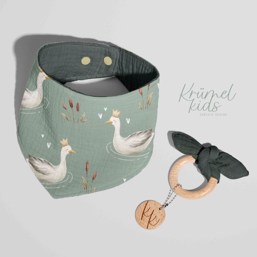 Baby muslin bandana mockup displayed flat on a light surface, featuring a swan pattern with a wooden teether beside it.