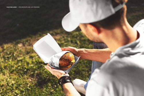 PSD mockup showing a wrapped burger inside an open box, held by a person outdoors in natural sunlight.