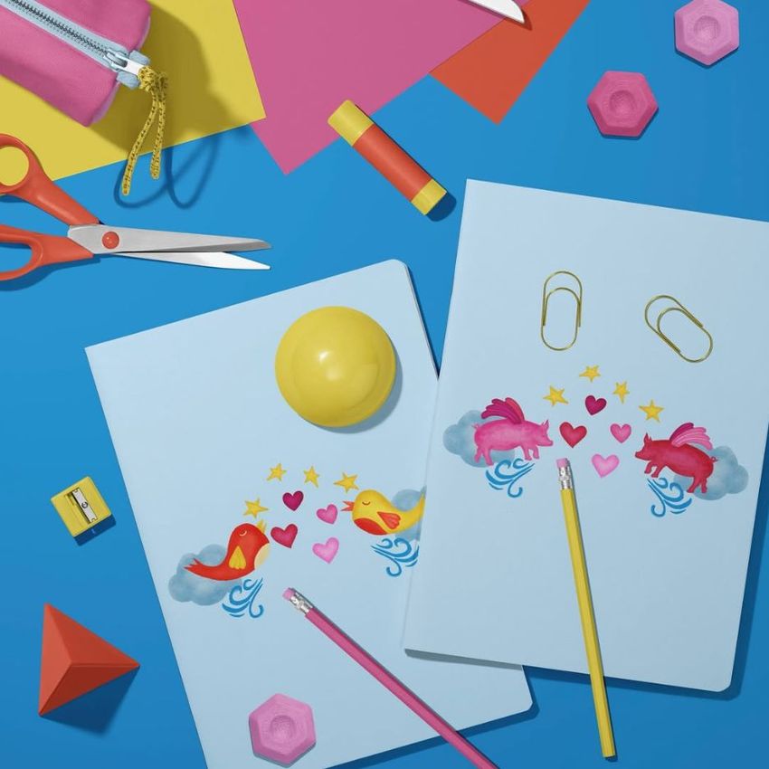 Stationery mockup featuring two classic A4 notebooks with colorful flying pig illustrations, pencils, and office supplies on a blue surface.