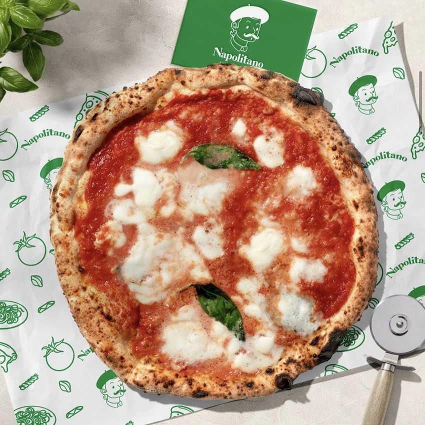Pizza mockup on food safe paper with green illustrated pattern, placed on a light surface with a pizza cutter and basil leaves.