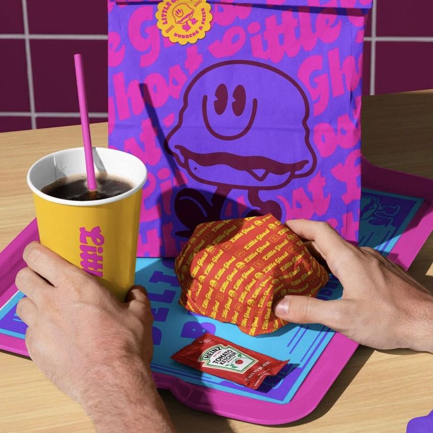 Burger restaurant mockup featuring a purple paper bag with ghost graphics, yellow drink cup, wrapped burger, and condiment packet.