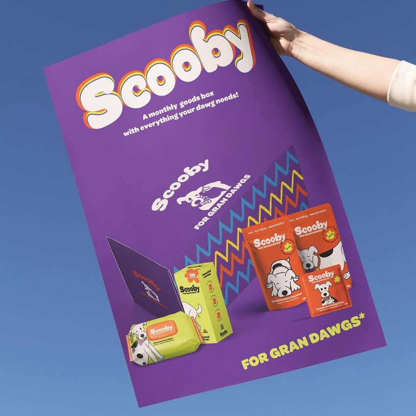 Hand holding a large purple poster mockup with colorful packaging designs, bold Scooby text, and illustrated dog products against a blue sky.