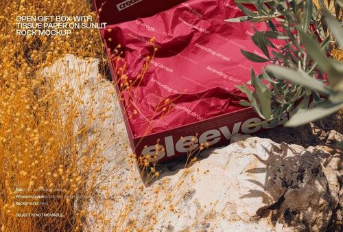 Box mockup with red tissue paper, displaying branding drops text, set on sunlit rocks surrounded by dry plants.