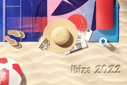 Beach towel mockup on sand with colorful geometric pattern, straw hat, sunglasses, flip-flops, and beach ball nearby.