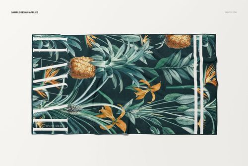 Top view of a beach towel mockup featuring green foliage, pineapples, and orange flowers on a dark background.