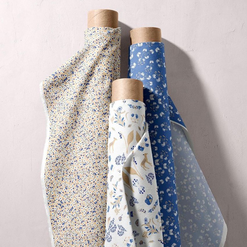 Fabric rolls mockup showing three upright rolls with floral and botanical patterns in blue, beige, and white tones against a light wall.