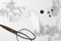 Mockup scene featuring wide cotton ribbon, metal scissors, and buttons arranged on a plain white textile, top perspective.
