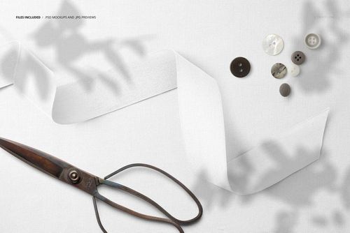 Mockup scene featuring wide cotton ribbon, metal scissors, and buttons arranged on a plain white textile, top perspective.