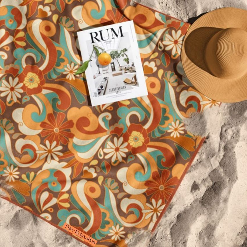 Beach towel mockup with a retro floral pattern in orange, teal, and cream, shown on sand with a magazine and straw hat.