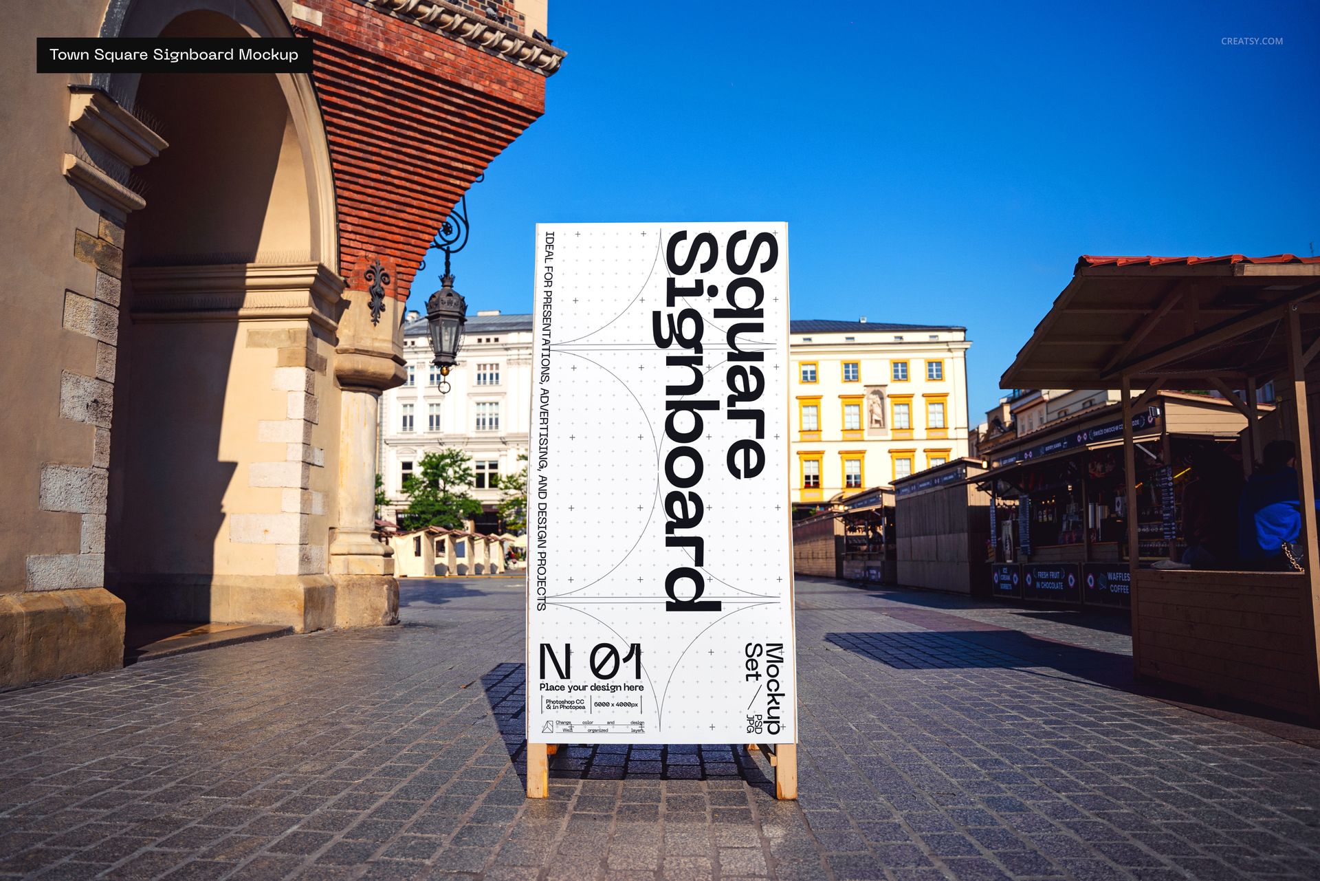Large square signboard mockup with black and white design standing in a sunny outdoor town square with historic buildings.