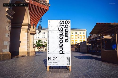 Large square signboard mockup with black and white design standing in a sunny outdoor town square with historic buildings.