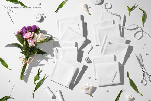 Jersey fabric mockup featuring folded white samples, fresh flowers, green leaves, and sewing items viewed from above.