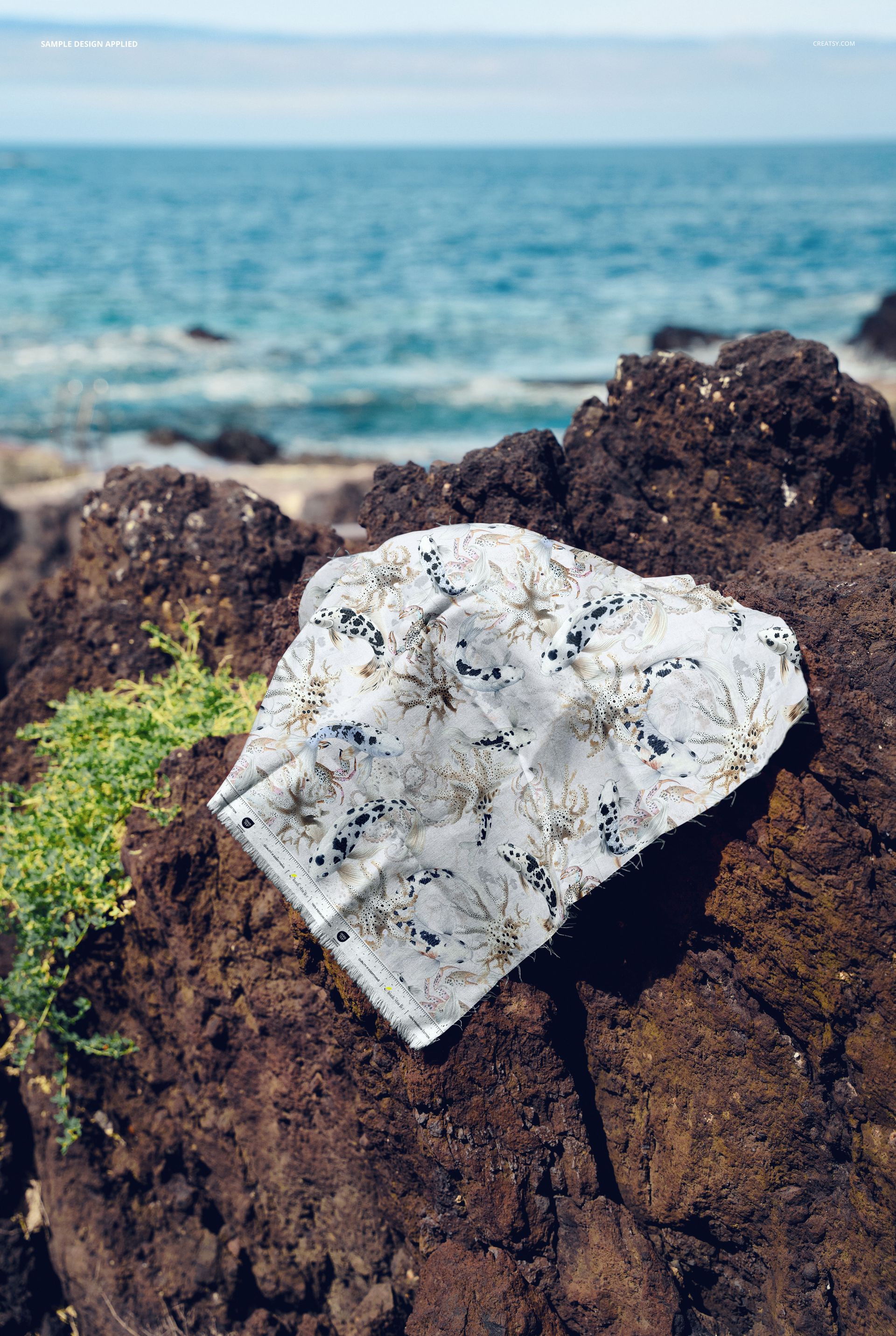 Floral patterned cotton fabric swatch mockup displayed on rugged rocks beside the sea, natural daylight scene.