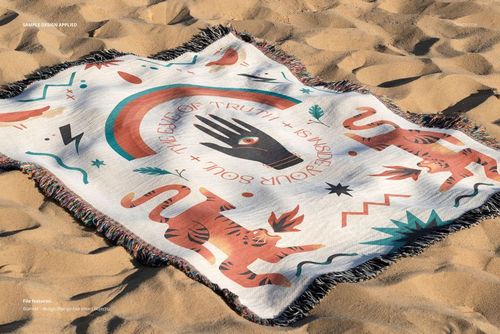 Jacquard fringed throw blanket mockup with geometric and hand motifs, laid flat on sand in natural light.
