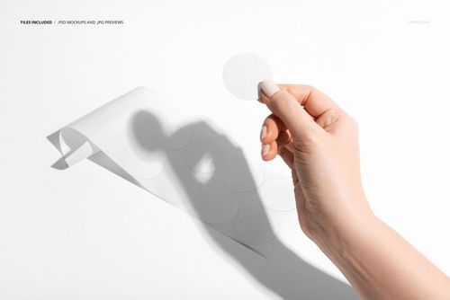 Round sticker being held by fingers next to a sheet of blank circle labels, photographed in bright lighting for a mockup.