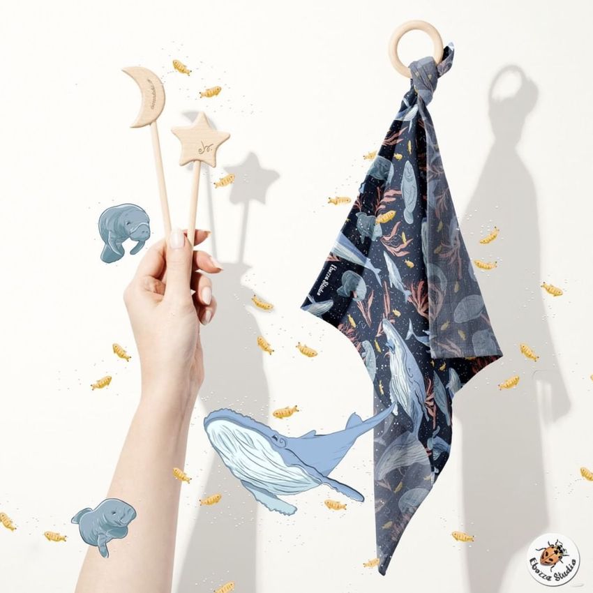 Muslin cloth wooden teether mockup featuring a patterned dark blue cloth with ocean animals, a wooden ring, and wooden wands held by a hand.