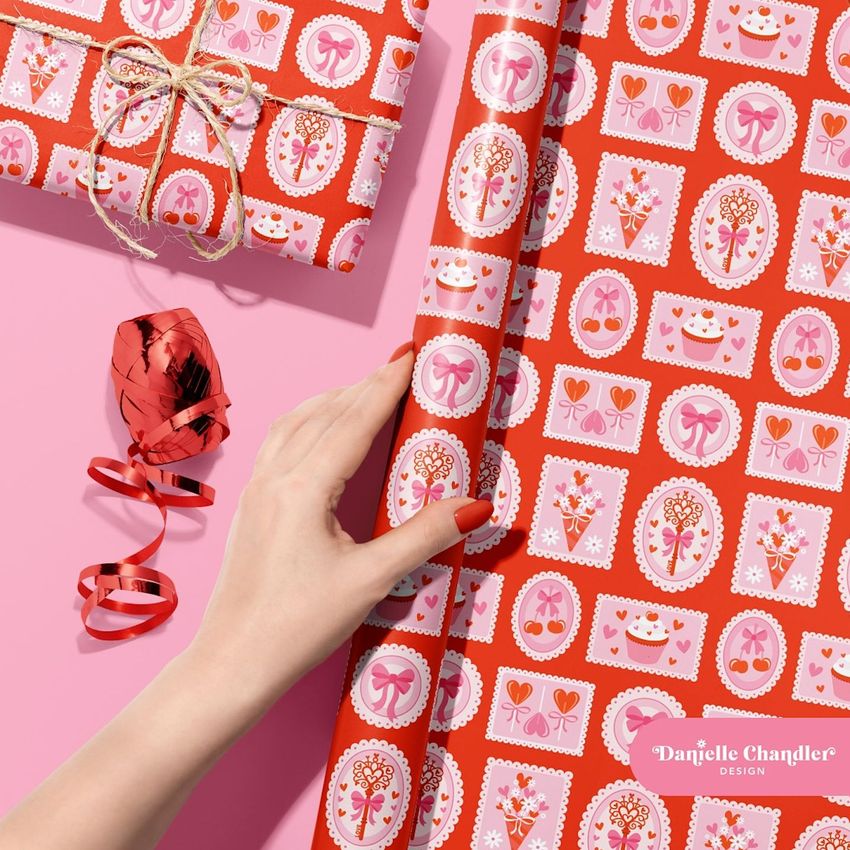 Gift wrapping paper mockup featuring a hand holding a roll of red patterned paper with pink and white motifs on a pink surface.