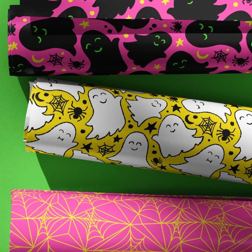 Three Halloween wrapping paper rolls with ghost, spider, and web patterns in black, pink, yellow, and white on a green surface.