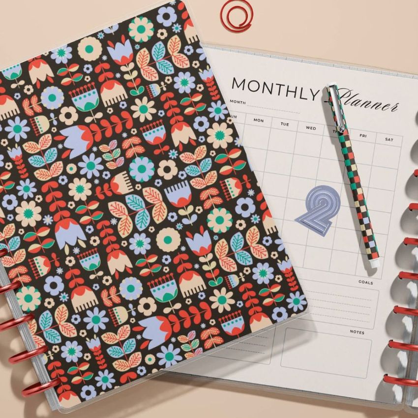 Spiral planner mockup showing a closed floral cover next to an open monthly planner, with a striped pen and red rings visible.
