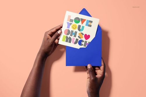 Square folded greeting card mockup featuring playful, colorful typography, held over a blue envelope on a light peach surface.