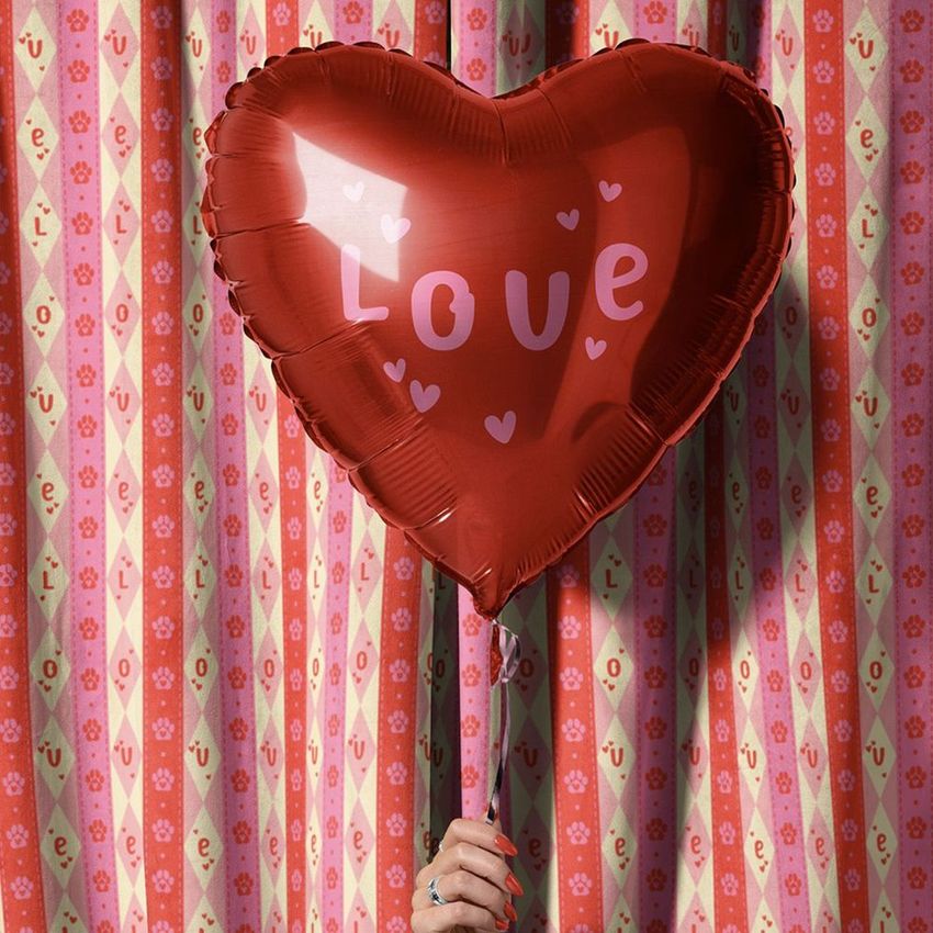 Hand holding a red heart-shaped balloon with the word 'Love' and small hearts, in front of a pink velvet curtain mockup.