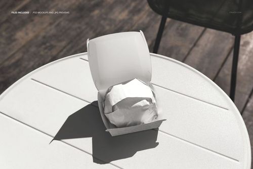 PSD mockup showing a wrapped burger inside a white takeaway box on a sunlit round table in an outdoor setting.