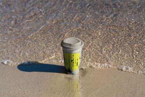 Single paper coffee cup mockup with a gray lid placed on sandy beach, water gently approaching the cup.