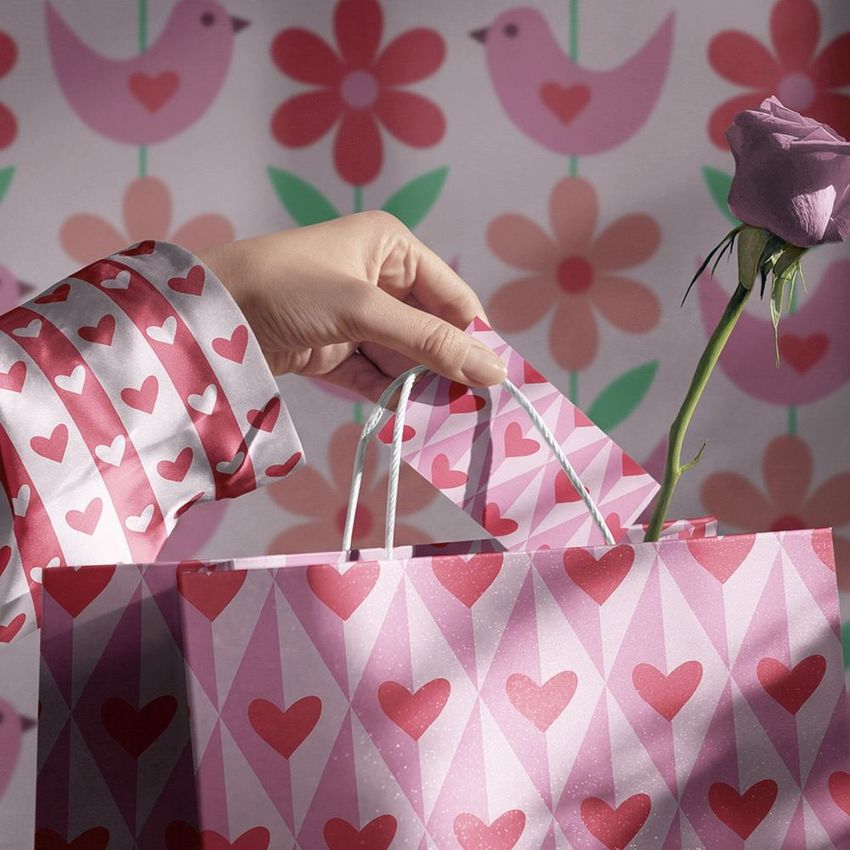 Gift bag mockup showing a hand adding a card to a heart-patterned bag, with a rose and bird and flower wallpaper behind.