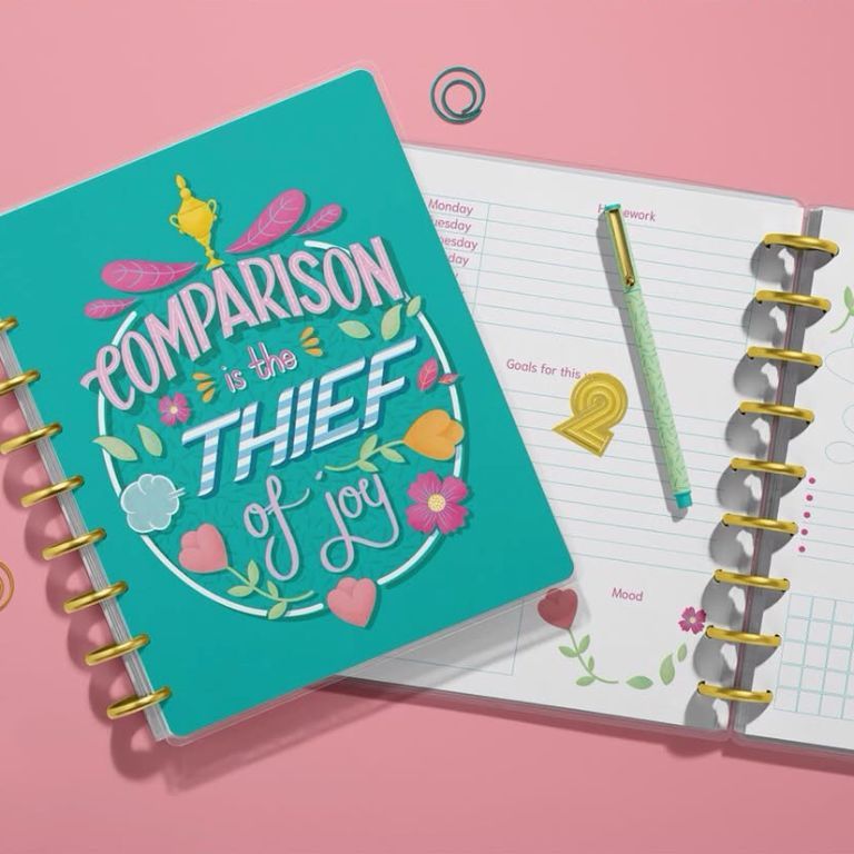 Top view of two spiral planners on a pink surface, one closed with colorful text and floral graphics, one open with lined pages.