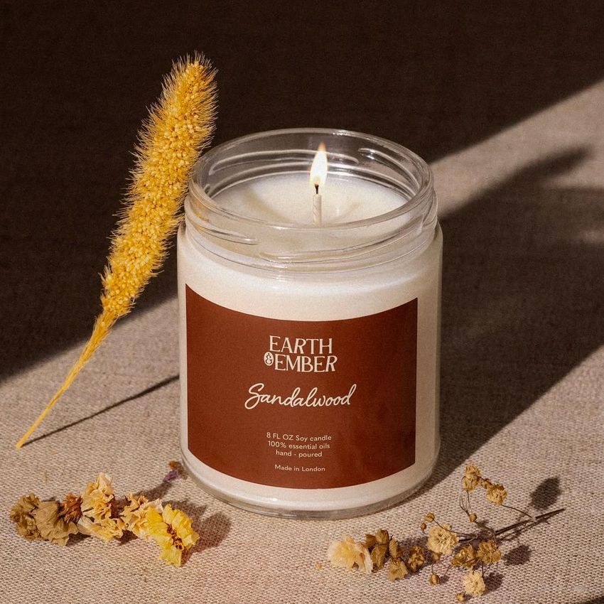 Scented soy candle mockup featuring a glass jar with a brown label, lit wick, and dried flowers on a neutral fabric background.