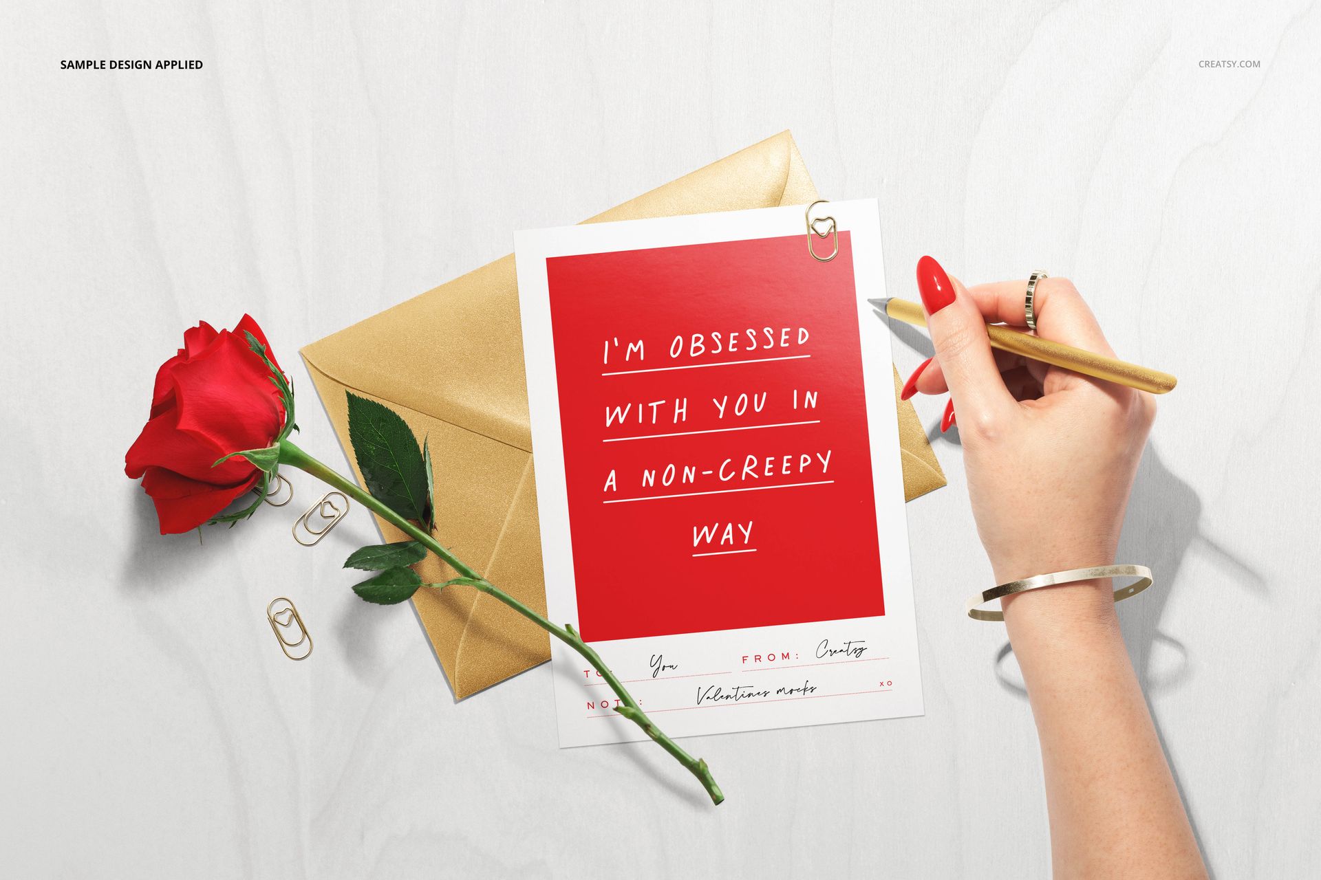 Top view of a Valentine's stationery mockup featuring a red card, gold envelope, rose, and hand holding a pencil.