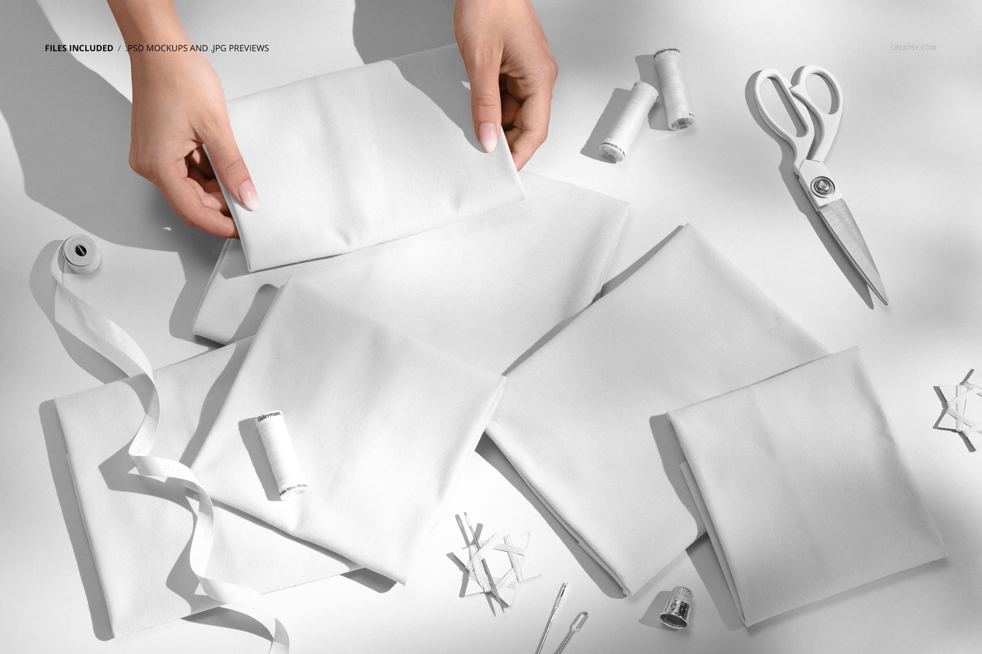 Hands arranging folded jersey fabric mockup, with spools of thread, ribbon, pins, and scissors on a white tabletop.