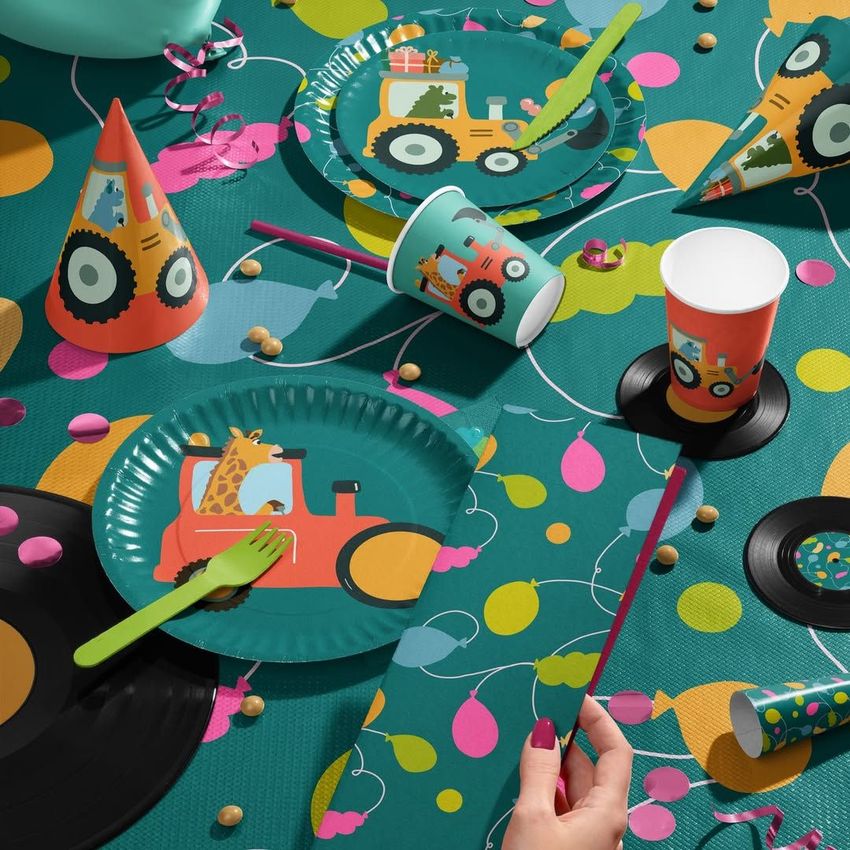 Party table with disposable tableware mockup featuring plates, cups, and hats decorated with colorful tractor illustrations.