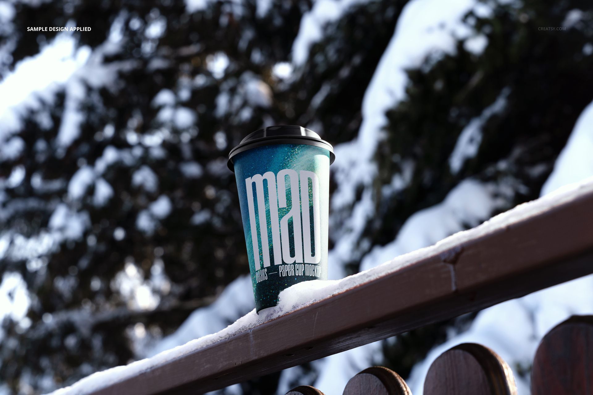 Winter paper cup mockup featuring a blue design and black lid, set on a snowy wooden fence with trees in background.