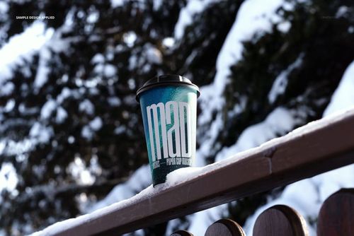 Winter paper cup mockup featuring a blue design and black lid, set on a snowy wooden fence with trees in background.