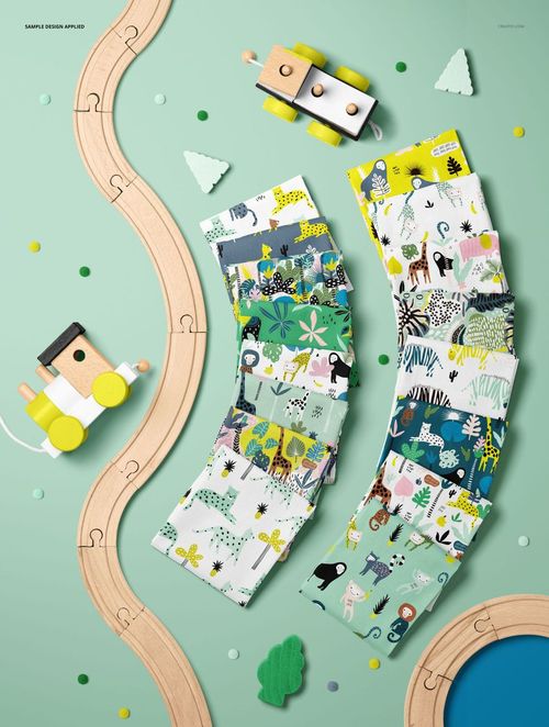 PSD mockup of patterned cotton fabric swatches with jungle animals, placed near wooden train tracks and toy trees on a light green table.