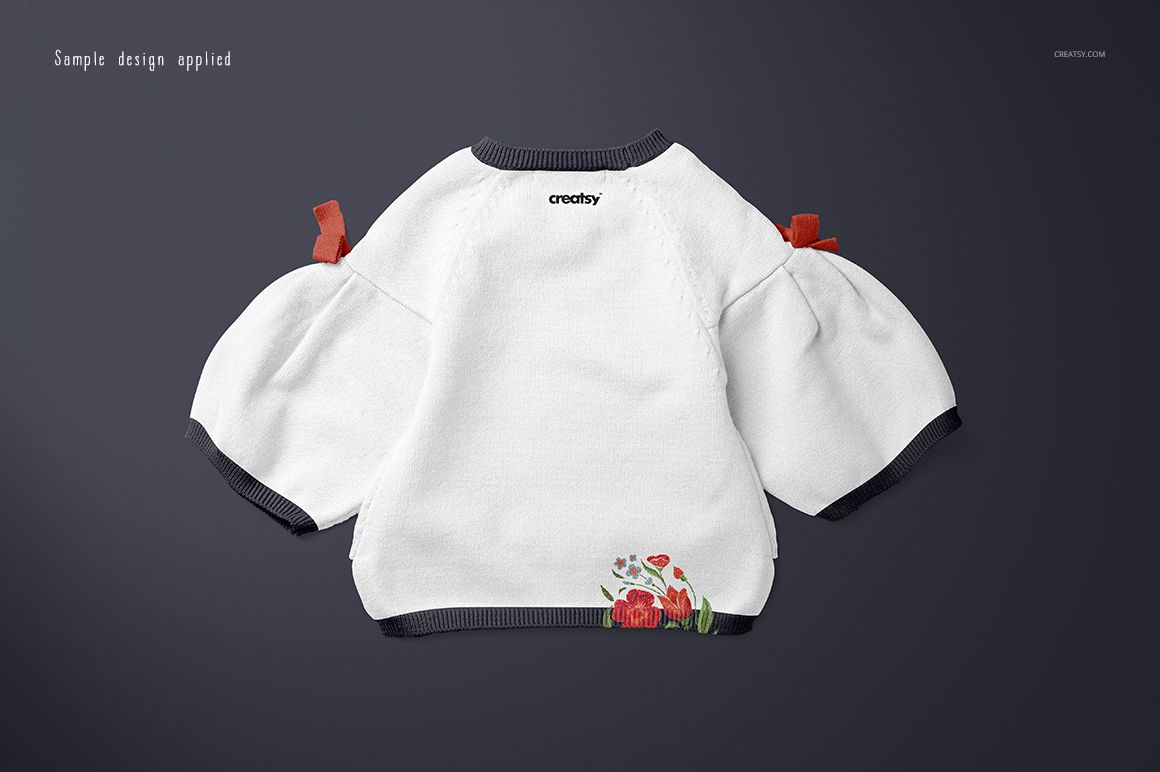 White baby sweater with dark cuffs and floral embroidery on the back, shown in a flat lay mockup on a dark background.
