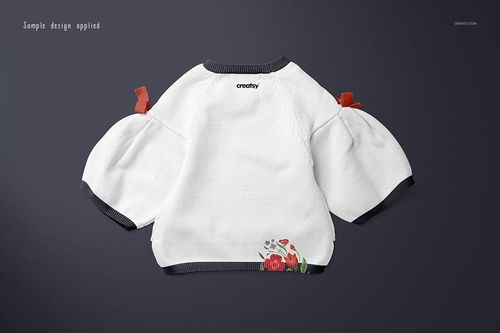White baby sweater with dark cuffs and floral embroidery on the back, shown in a flat lay mockup on a dark background.