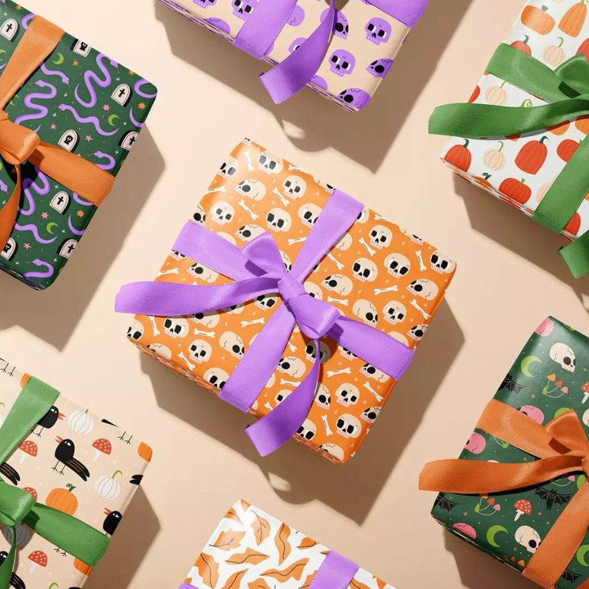 Gift box mockup featuring several wrapped boxes with Halloween-themed patterns, including skulls, pumpkins, and snakes, arranged on a beige surface.