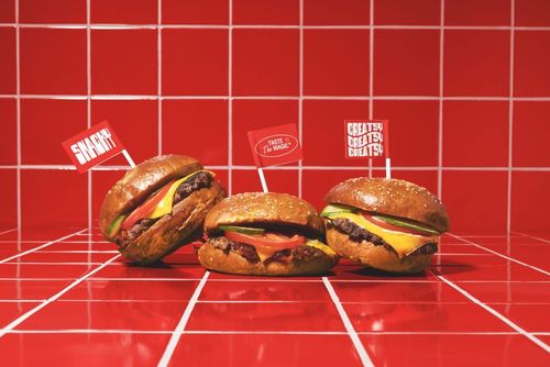 PSD mockup showing three cheeseburgers with toppings and branded flags, arranged on a red tiled countertop.