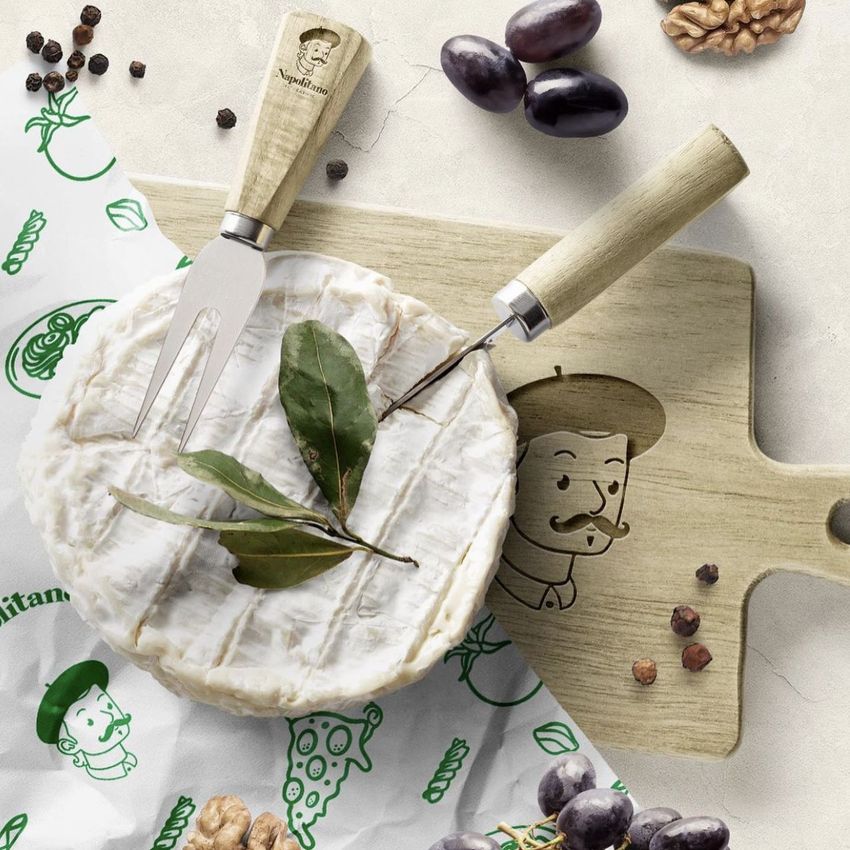 Camembert cheese on a wooden board mockup with cheese knives, olives, walnuts, and peppercorns on a light surface.