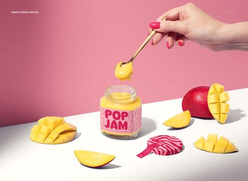 Front view of a mango jam jar mockup with a pink label, surrounded by fresh mango slices and a hand holding a spoon.