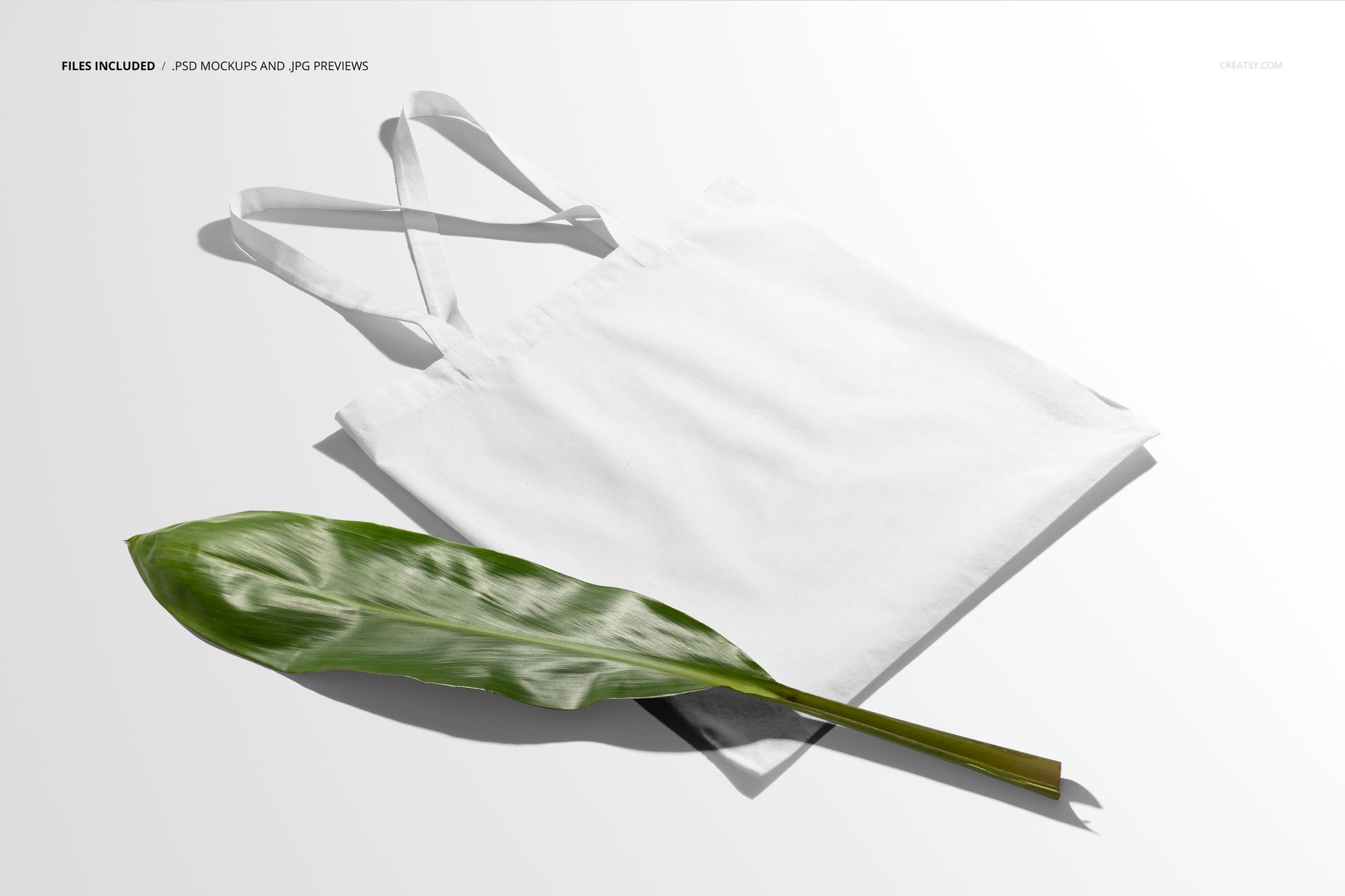 White tote bag mockup displayed on a light surface, viewed from above with a green leaf placed in front of the bag.