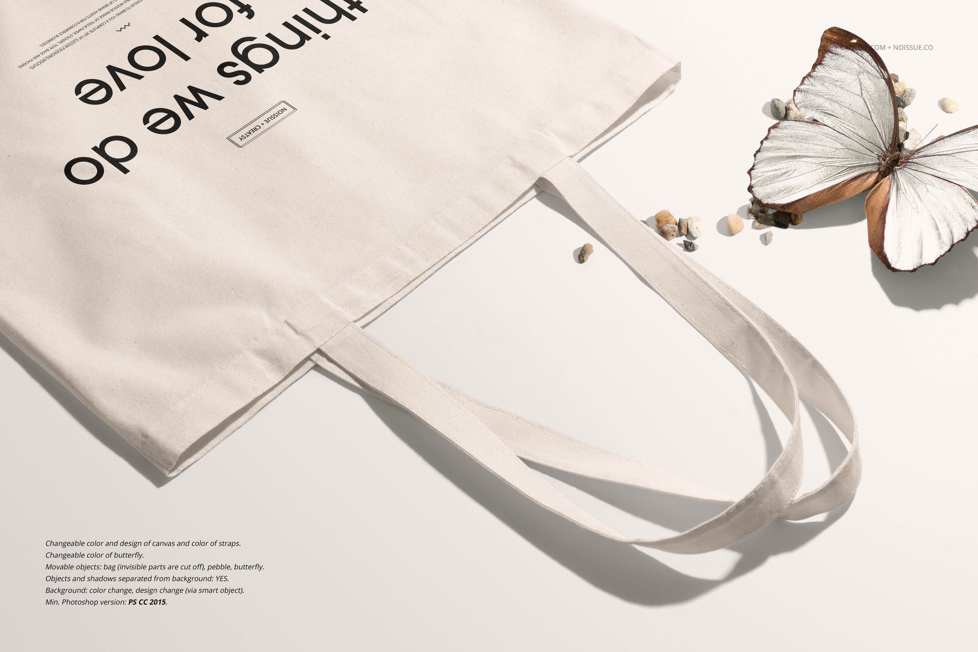 Front view of a beige tote bag mockup with black text, placed on a light surface with a butterfly and small pebbles.