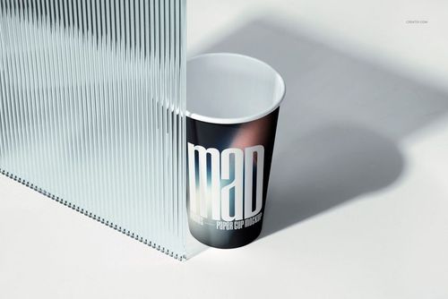 PSD mockup of a paper cup with a modern graphic print, casting a shadow beside a textured glass panel on a clean surface.