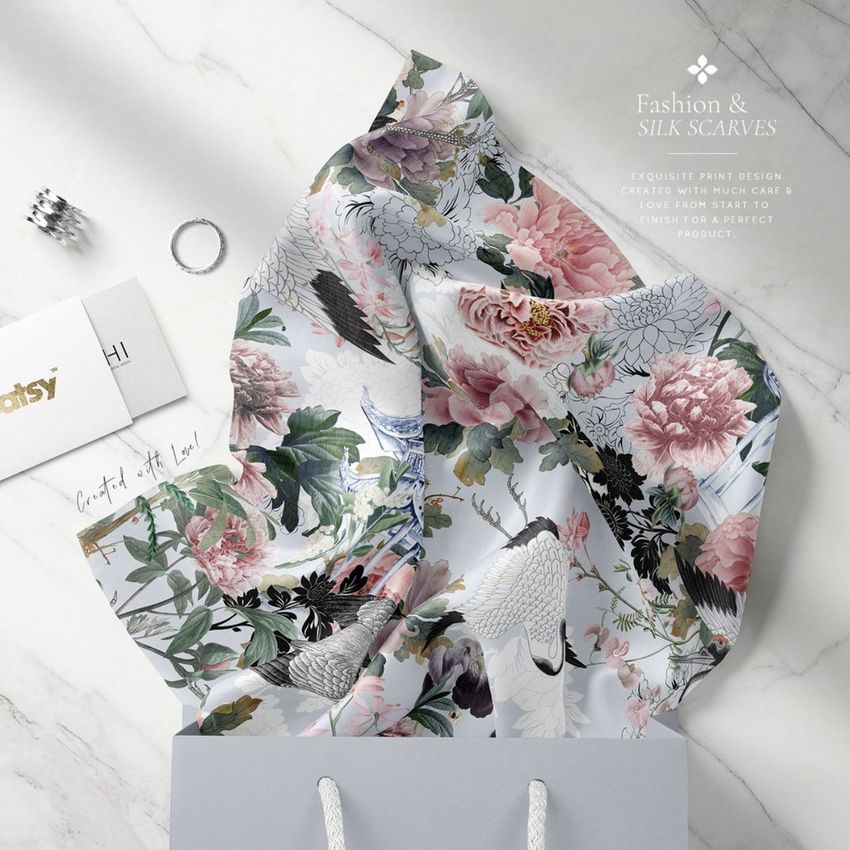 Silk scarf mockup featuring a floral and crane pattern in soft pastel colors, draped over the edge of a gray shopping bag on a light surface.
