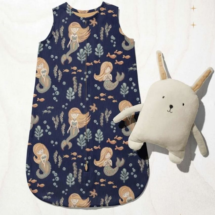 Baby wearable blanket mockup featuring a navy blue fabric with whimsical mermaid and fish illustrations, shown beside a plush rabbit toy.