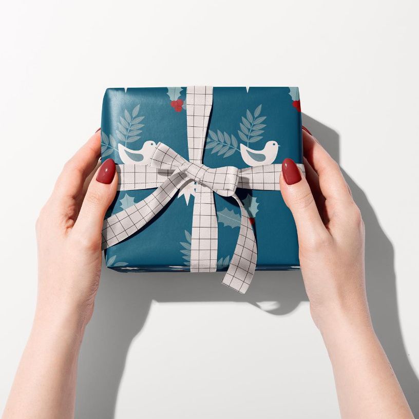 Gift box mockup wrapped in blue paper with white bird and leaf pattern, tied with a grid-patterned ribbon, held by two hands.